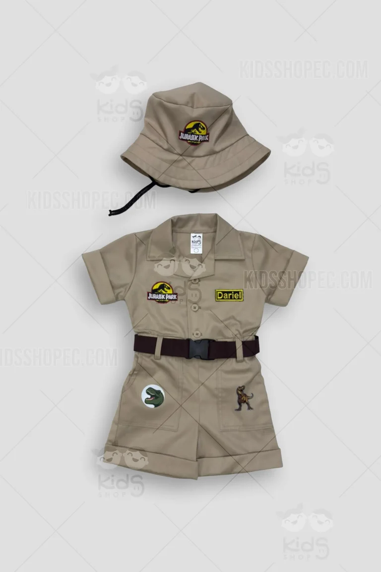 Fotografía de un traje infantil de explorador estilo safari en color caqui (beige). Consta de una camisa de manga corta con botones frontales, pantalón corto con bolsillos y un cinturón marrón con hebilla plástica negra. Incluye parches decorativos temáticos de dinosaurios (T-Rex) en el pantalón y un parche estilo parque jurásico en el pecho y en el sombrero tipo pescador (bucket hat) a juego. En el lado izquierdo del pecho lleva un parche amarillo personalizado con el nombre "Dariel". Fondo gris neutro con marcas de agua de Kids Shop EC.