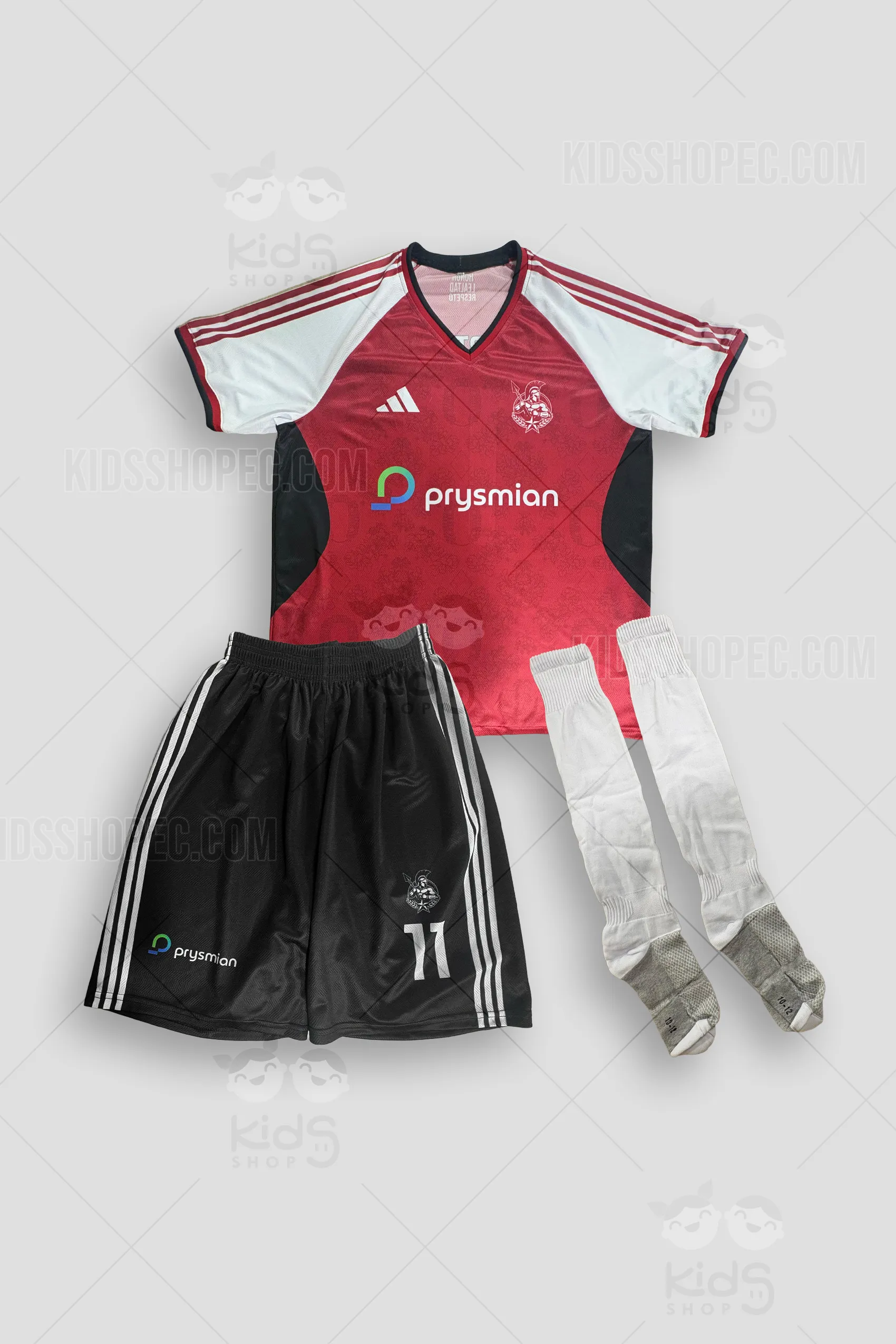 Fotografía de un uniforme de fútbol completo compuesto por tres piezas. La camiseta de cuello en "V" tiene un cuerpo de color rojo carmesí con un sutil patrón de escudos entrelazados en el fondo, paneles laterales negros y mangas blancas raglán con tres franjas rojas. Incluye el escudo de un guerrero espartano, logo del patrocinador "Prysmian" y etiquetas de Winma Sports. La pantaloneta es negra con tres franjas blancas laterales, llevando el número 11 y los mismos logos sublimados. Se incluyen un par de medias deportivas blancas con refuerzos grises en talón y punta. Fondo gris neutro con marcas de agua de Kids Shop EC.