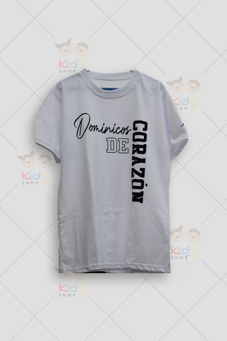 Fotografía de una camiseta básica blanca de manga corta y cuello redondo. En la parte frontal lleva un estampado tipográfico asimétrico en color negro que dice "Dominicos DE CORAZÓN". La palabra "Dominicos" está en una fuente cursiva elegante, "DE" en letras de contorno tipo universitario, y "CORAZÓN" está escrito verticalmente en letras mayúsculas en negrita. La prenda se exhibe plana sobre un fondo gris neutro con marcas de agua de Kids Shop EC.