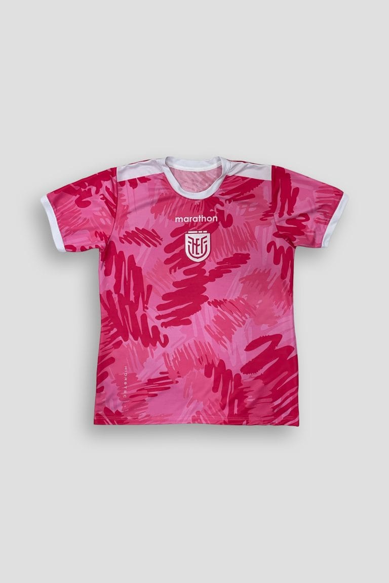 Fotografías de estudio de una camiseta deportiva de manga corta destacada por su vibrante color rosado y un patrón abstracto de trazos dinámicos en tonos magenta y rosa claro. Presenta cuello redondo y bordes de las mangas en color blanco contrastante. En el centro del pecho lleva estampado en color blanco un escudo deportivo representativo del fútbol ecuatoriano junto a letras de estilo deportivo. Las imágenes muestran la vista frontal completa, un primer plano de los detalles del pecho y la espalda limpia sobre un fondo gris claro.