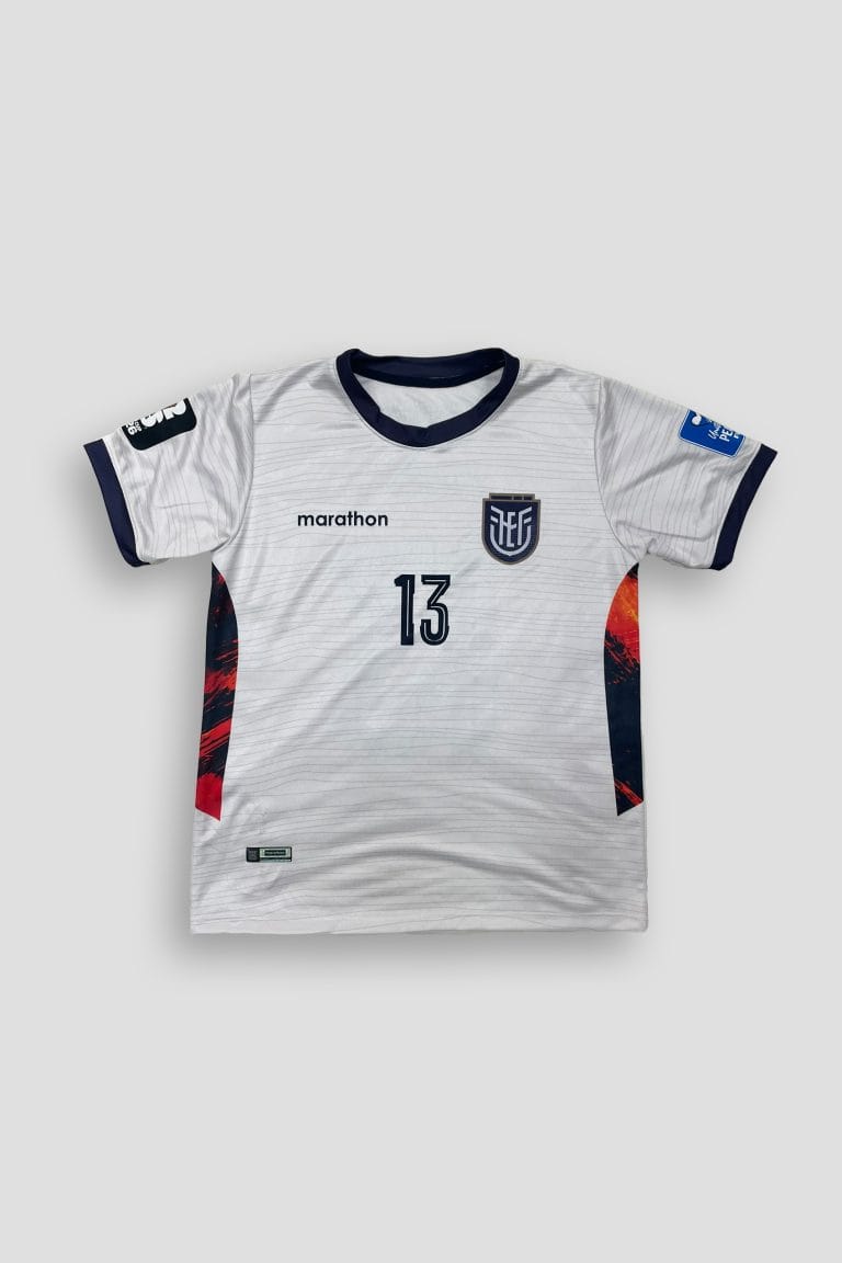 Fotografías de estudio de una camiseta deportiva de fútbol de manga corta, con base de color blanco texturizada con finas líneas horizontales grises. Presenta cuello redondo y puños en color azul marino, y paneles laterales con gráficos abstractos en rojo, azul y amarillo. En el pecho izquierdo lleva estampado un escudo representativo deportivo en azul marino y gris, junto al número 13 frontal. En la espalda destaca el texto 'E. VALENCIA' impreso sobre un número 13 grande en azul marino, además de una frase pequeña en la parte superior trasera. Fondo gris claro.